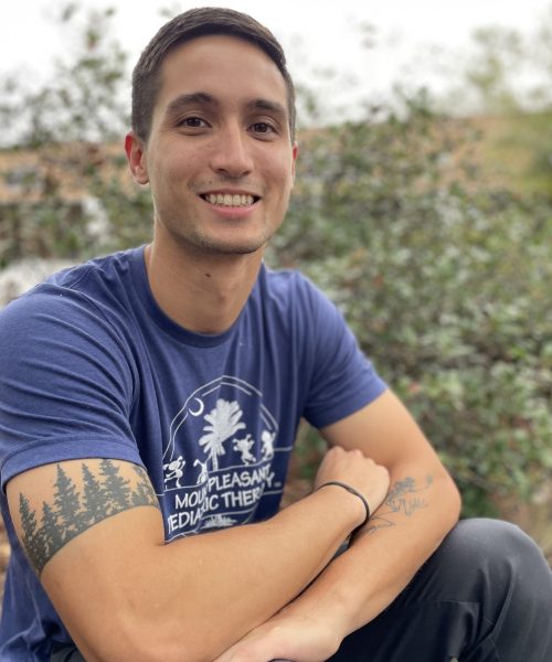 Man smiling outdoors with forest tattoo visible.