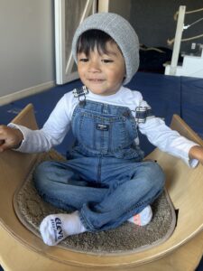 Smiling toddler sitting cross-legged in casual denim overalls and a beanie.