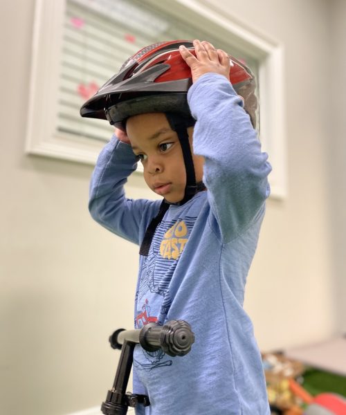 Child wearing helmet, holding scooter indoors.